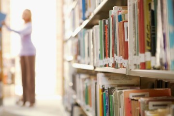 Je ziet een bibliotheekgang met boeken. Vaag op de achtergrond staat een persoon die een boek uit het rek neemt.