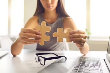 Je ziet een vrouw met twee puzzelstukken in hout die ze naar elkaar toe brengt. Op de voorgrond is ligt er een bril en een laptop op tafel.